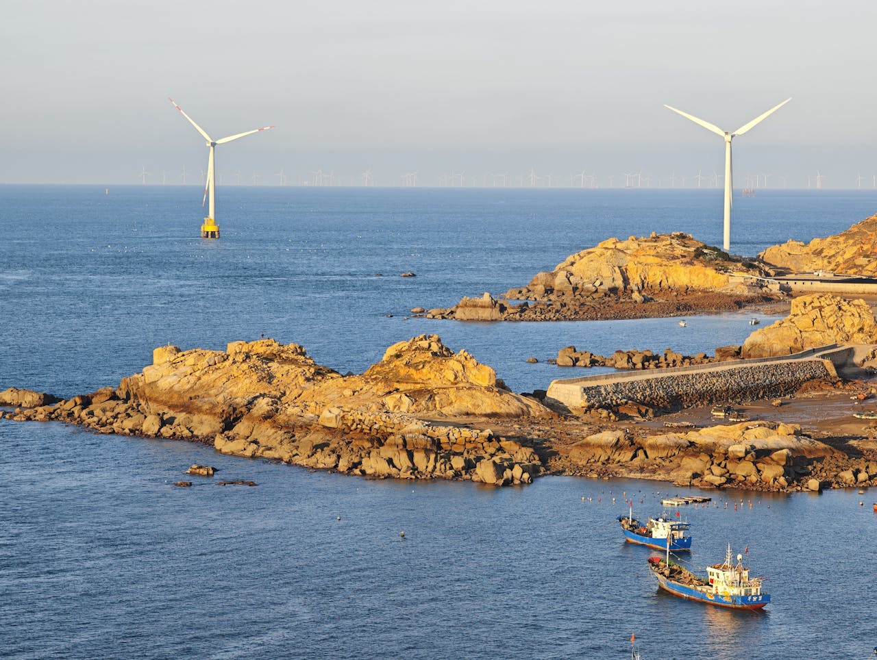 Scenic view of offshore wind turbines near a rocky coastline with boats in tranquil waters.